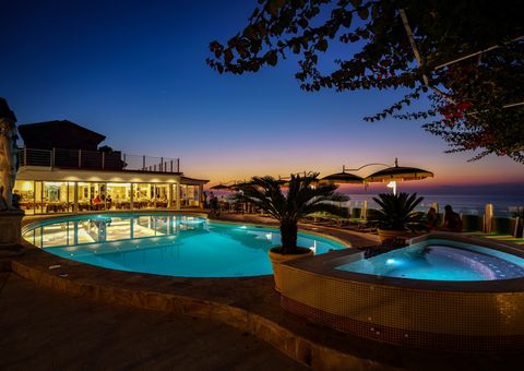 Stimmungsvolle Abendkulisse am beleuchteten Pool mit Whirlpool, gemütlichen Sitzplätzen und Blick auf den farbenprächtigen Sonnenuntergang über dem Meer.