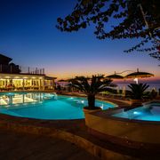 Stimmungsvolle Abendkulisse am beleuchteten Pool mit Whirlpool, gemütlichen Sitzplätzen und Blick auf den farbenprächtigen Sonnenuntergang über dem Meer.