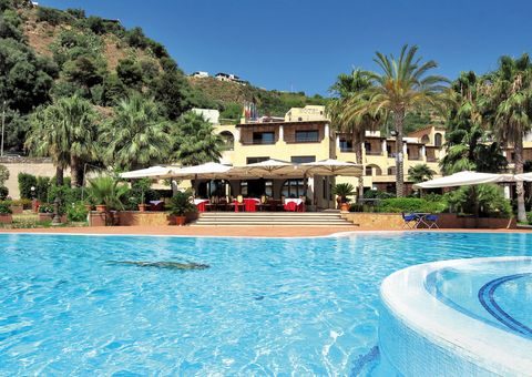 Großer Außenpool vor einem mediterranen Hotelgebäude mit Sonnenterrasse, Palmen und grüner Hügellandschaft im Hintergrund – sonnige Urlaubsatmosphäre unter blauem Himmel.