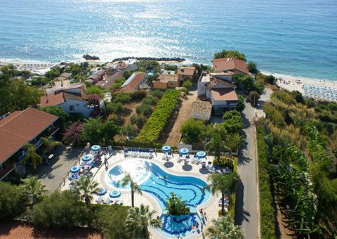 Luftaufnahme eines Hotels an der Küste mit geschwungenem Außenpool, umgeben von Palmen und Sonnenschirmen; im Hintergrund das tiefblaue Meer und ein Sandstrand mit Liegen.
