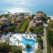 Luftaufnahme eines Hotels an der Küste mit geschwungenem Außenpool, umgeben von Palmen und Sonnenschirmen; im Hintergrund das tiefblaue Meer und ein Sandstrand mit Liegen.