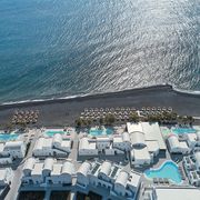 Luftaufnahme des Costa Grand Resort & Spa auf Santorin – elegante Hotelanlage direkt am schwarzen Lavastrand von Kamari mit mehreren Pools, weißen Gebäuden im Kykladenstil und herrlichem Blick auf das funkelnde Meer.