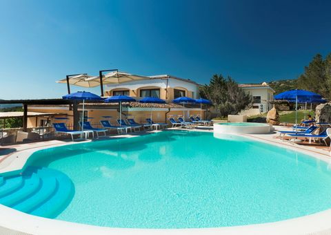 Einladender Außenpool mit türkisblauem Wasser, umgeben von blauen Sonnenschirmen und Liegestühlen vor einer mediterranen Hotelanlage unter klarem Himmel.