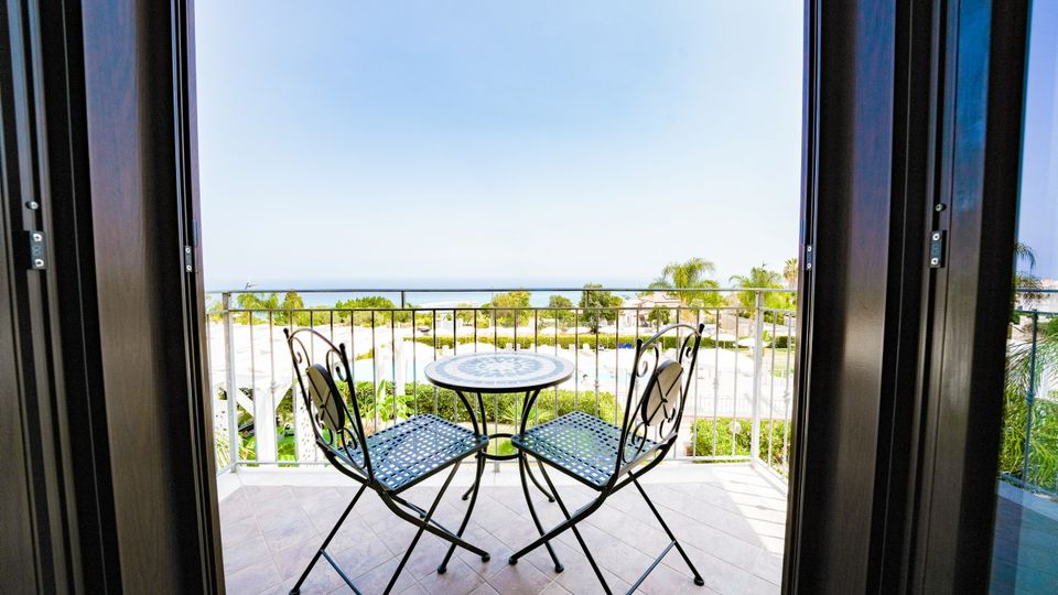 Balkon mit zwei Eisenstühlen und Tisch, Blick auf Poolanlage und das Meer in der Ferne.