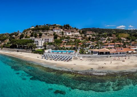 Panoramablick auf eine Hotelanlage direkt am klaren, türkisblauen Meer mit Liegestühlen am Sandstrand.