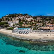 Panoramablick auf eine Hotelanlage direkt am klaren, türkisblauen Meer mit Liegestühlen am Sandstrand.