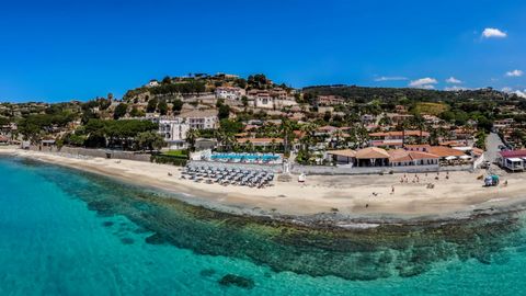 Panoramablick auf eine Hotelanlage direkt am klaren, türkisblauen Meer mit Liegestühlen am Sandstrand.