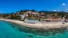 Panoramablick auf eine Hotelanlage direkt am klaren, türkisblauen Meer mit Liegestühlen am Sandstrand.