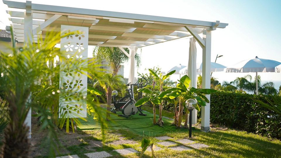 Überdachte Outdoor-Trainingsfläche mit Heimtrainer, umgeben von Palmen und tropischer Bepflanzung in gepflegter Gartenanlage.